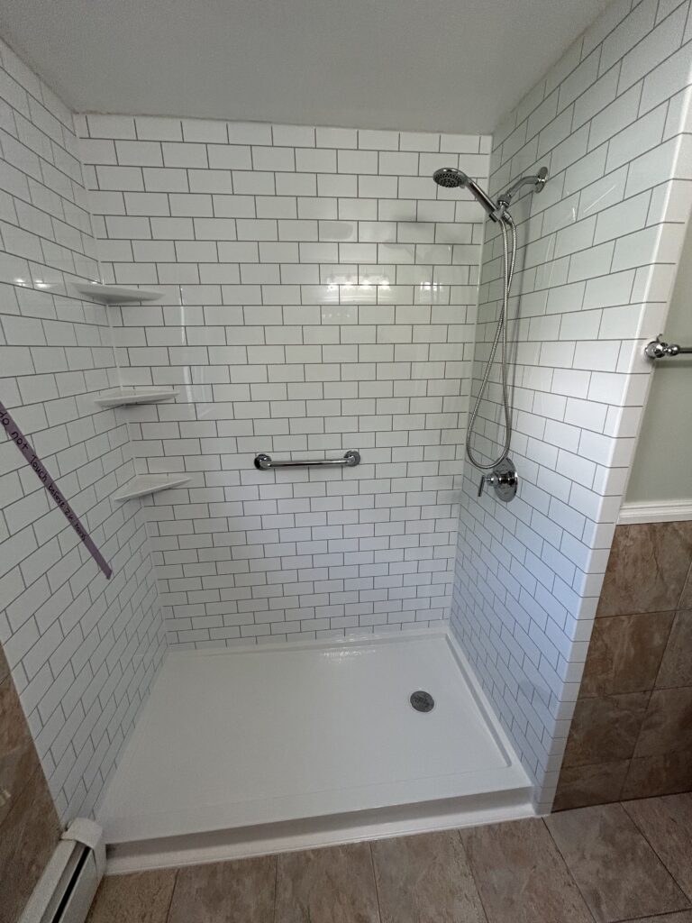 Shower conversion featuring white surround walls in a classic subway tile pattern with contrasting black grout. Includes a low-barrier white shower pan, ADA-compliant grab bar, built-in corner shelves, and a brushed-nickel rain showerhead with handheld sprayer.